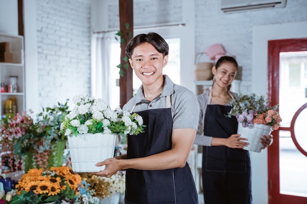 Florista jovem feliz vestindo avental, segurando balde de flores sorrindo, olhando para a câmera. trabalhando em uma floricultura com seu amigo atrás