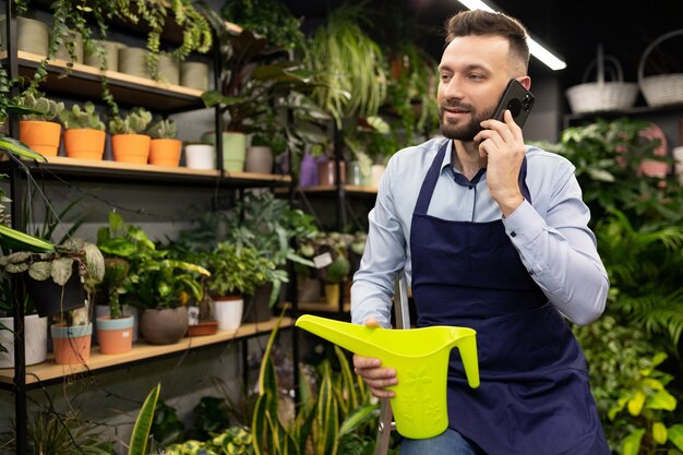 un florista masculino en una floristería con una lata de agua en las manos habla por teléfono en el contexto de las plantas en maceta.