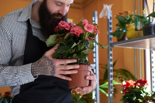 Florista masculino profesional con barba y tatuaje en la mano con uniforme sostiene la olla con un ramo de flores en la floristería.