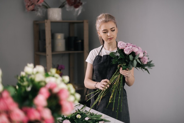 Florista profissional jovem vestindo avental fazendo arranjo floral de rosa fresca na mesa sobre fundo branco. conceito de trabalhar com flores, negócios florais.