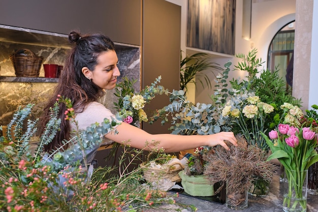 Florista sonriente en delantal mirando las flores en flor y arreglando un ramo en una tienda de flores moderna