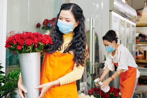 Floristería asiática joven en máscara médica mirando el cubo de rosas rojas frescas en sus manos