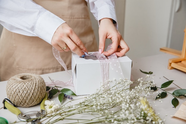 Una floristería decora una caja de regalo con flores y una cinta en un escritorio blanco, solo las manos están en el marco.