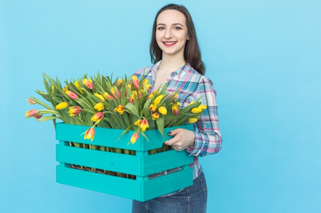 Floristería mujer caucásica feliz riendo y sosteniendo gran caja de tulipanes en superficie azul