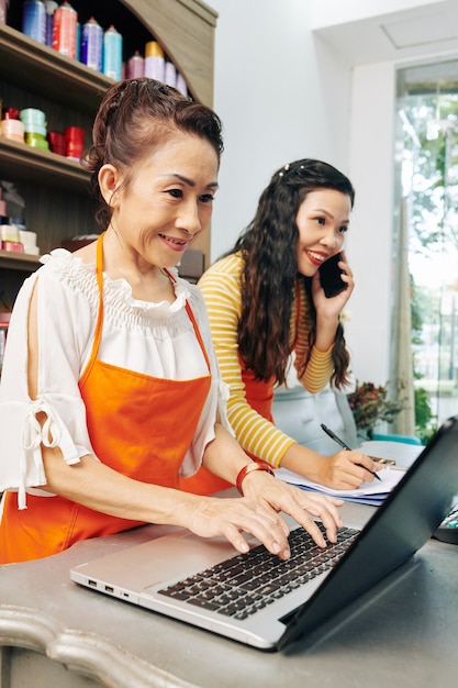 Foto floristería senior aceptando el pedido a través del formulario en el sitio web de la tienda de flores cuando su colega más joven hablando por teléfono con el cliente