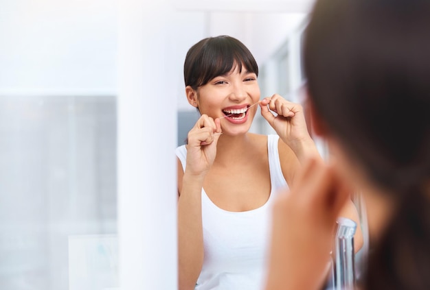 Flossing away Foto de una alegre y atractiva mujer joven que se pasa el hilo dental mientras mira su reflejo en un espejo en casa