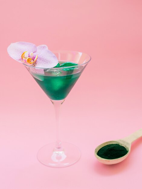 Flüssiges Spirulina-Grüngetränk mit Phalaenopsis im Cocktailglas und Spirulina-Pillen auf rosafarbenem Hintergrund