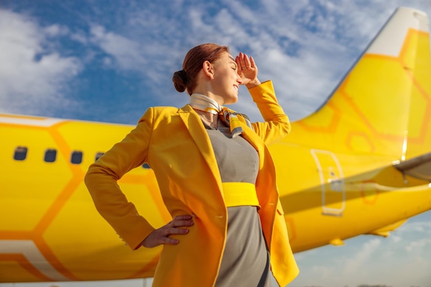 Flugbegleiterin in gelber Jacke, die mit der Hand die Augen vor der Sonne bedeckt und lächelt, während sie draußen auf dem Flugplatz steht