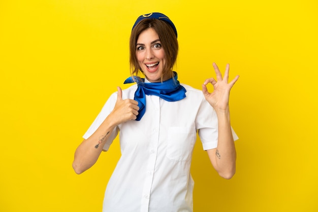 Flugzeug-Stewardess-Frau isoliert auf gelbem Hintergrund mit OK-Zeichen und Daumen hoch