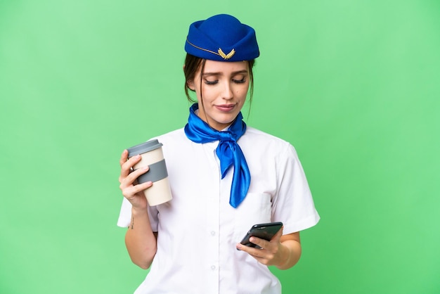 Flugzeugstewardess über isoliertem Chroma-Key-Hintergrund mit Kaffee zum Mitnehmen und einem Handy