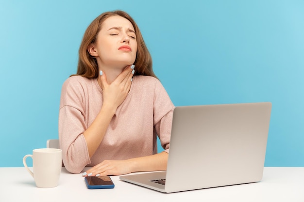Flusick joven empleada sentada en el lugar de trabajo con una computadora portátil y sosteniendo el cuello doloroso que sufre dolor de garganta amígdalas inflamadas síntomas de influenza estudio interior tiro aislado sobre fondo azul
