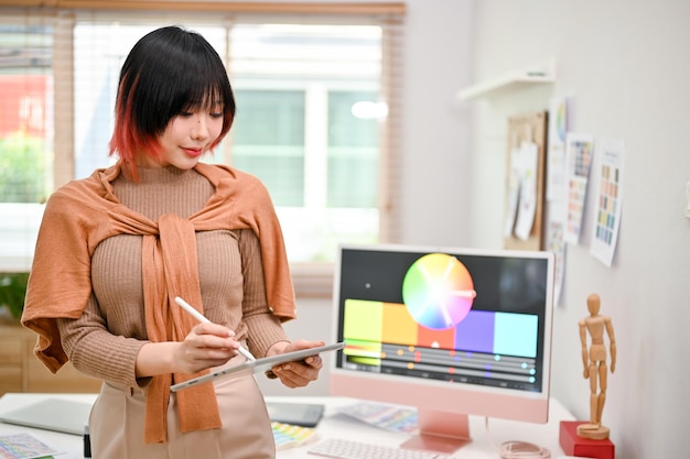Focada jovem designer gráfica asiática fica em seu estúdio e usa seu tablet digital