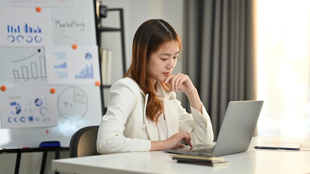 Focada jovem economista feminina analisando o mercado financeiro na tela do computador portátil em sua estação de trabalho