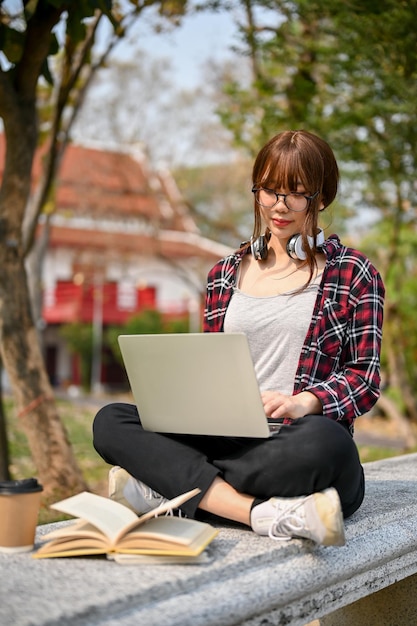 Focada jovem estudante universitária asiática usando laptop em um banco no parque do campus