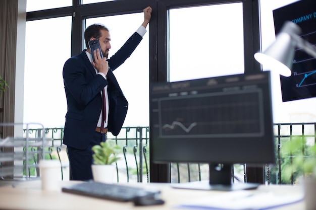 Focado jovem empresário tendo uma conversa telefônica e olhando pela janela. Jovem empresário em seu escritório.