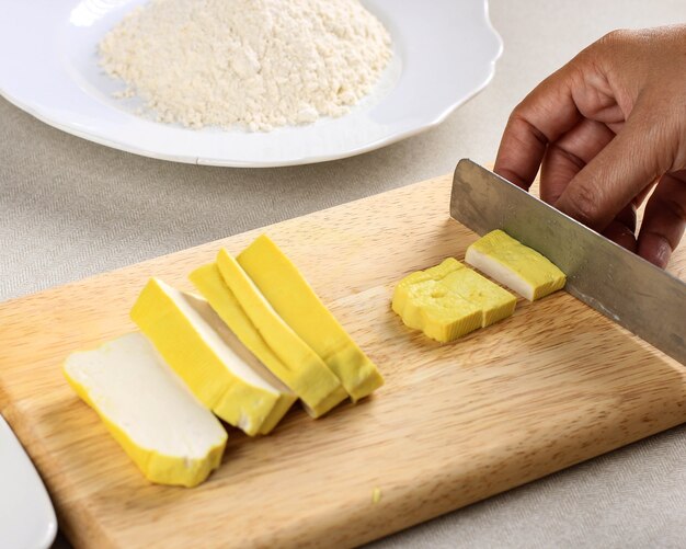 Foco Selecionado Preparando Refeição Vegana. Mão de uma chef feminina asiática cortando tofu amarelo com a faca na mesa de madeira