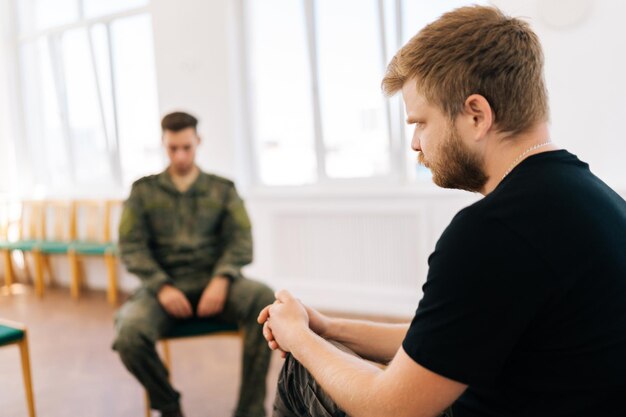 Foco selectivo de un militar caucásico deprimido en uniforme de camuflaje con TEPT hablando teniendo sesión de terapia de grupo con un especialista sentado en círculo