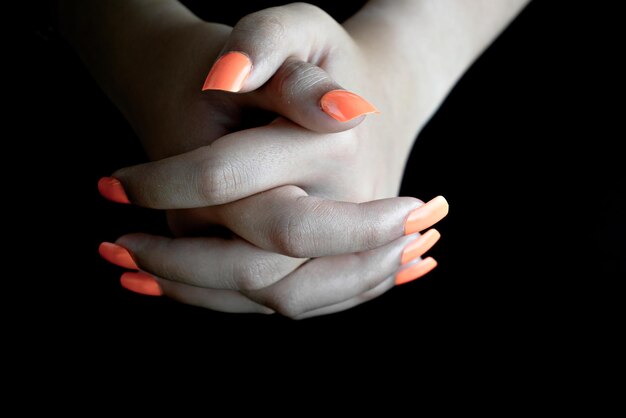 Foco seletivo de mãos femininas entrelaçadas com unhas postiças laranja, sobre fundo preto. manicure.