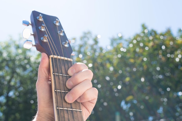 Foco seletivo de uma mão segurando um violão ao ar livre com o céu e a vegetação ao fundo