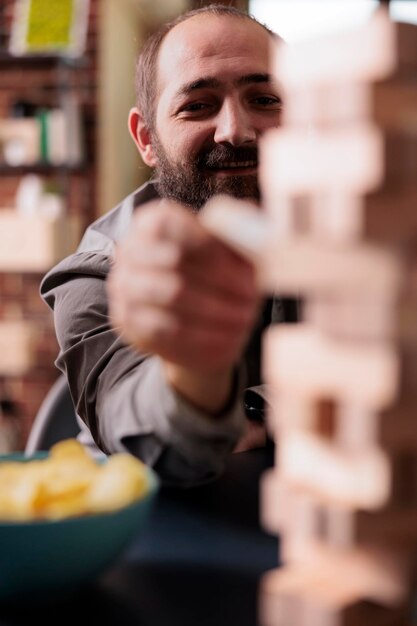 Foco seletivo do homem sorridente puxando o cubo de madeira da estrutura da torre de blocos. Pessoa feliz desfrutando da companhia de amigos enquanto jogava jogos de sociedade juntos em casa na sala de estar.