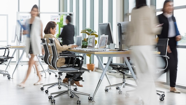 Foco seletivo em uma mulher de negócios sentada e trabalhando em laptops na mesa de conferência com outras mulheres de negócios embaçadas andando com pressa no escritório com várias janelas de vidro.