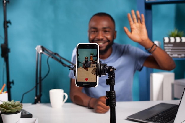 Foco seletivo no influenciador de mídia social de gravação de celular acenando olá sentado na mesa com microfone profissional. Closeup de configuração de vlog ao vivo com smarthone no vlogger de filmagem de estande.