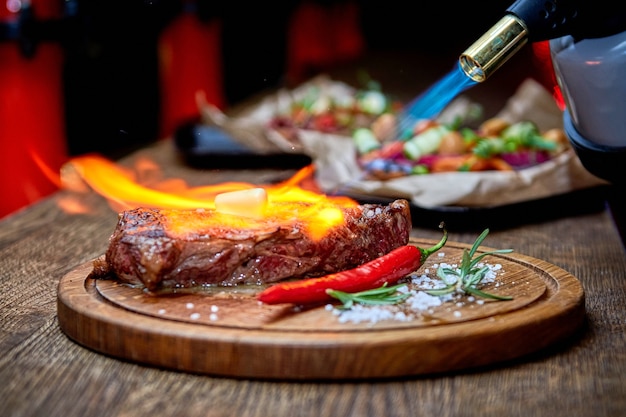 Foco suave. Entrecosto de carne Carne de bife grelhada com chamas de fogo na tábua de corte de madeira com ramo de alecrim, pimenta e sal. Chef master cozinhando um delicioso churrasco. Manteiga derretida