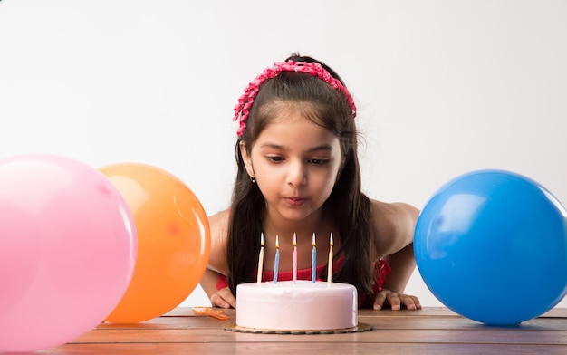 Fofa adorável garotinha indiana ou asiática comemorando aniversário segurando um bolo de morango e soprando velas na mesa ou em pé isolado sobre um fundo branco ou vermelho