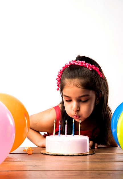 Fofa adorável garotinha indiana ou asiática comemorando aniversário segurando um bolo de morango e soprando velas na mesa ou em pé isolado sobre um fundo branco ou vermelho