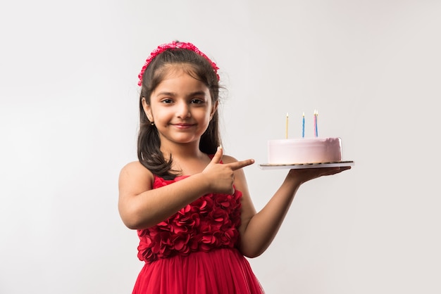 Fofa adorável garotinha indiana ou asiática comemorando aniversário segurando um bolo de morango e soprando velas na mesa ou em pé isolado sobre um fundo branco ou vermelho