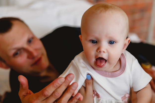 Fofa e feliz menina de 6 meses de olhos azuis e vestida de branco brincando na cama com o pai do homem