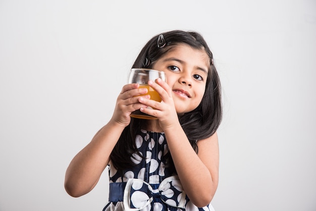 Fofinho menina indiana ou asiática brincalhona bebendo manga fresca ou suco de laranja ou bebida gelada ou bebida em um copo, isolado sobre fundo branco