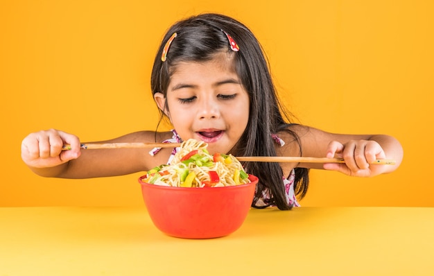 Fofinho menina indiana ou asiática comendo macarrão chinês saboroso com garfo ou pauzinhos, isolado sobre um fundo colorido