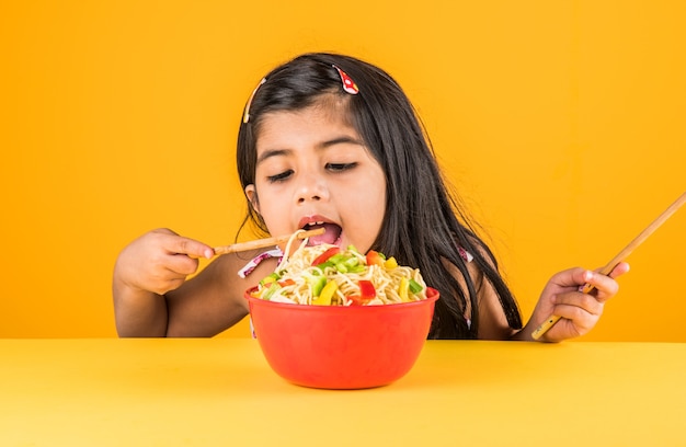 Fofinho menina indiana ou asiática comendo macarrão chinês saboroso com garfo ou pauzinhos, isolado sobre um fundo colorido