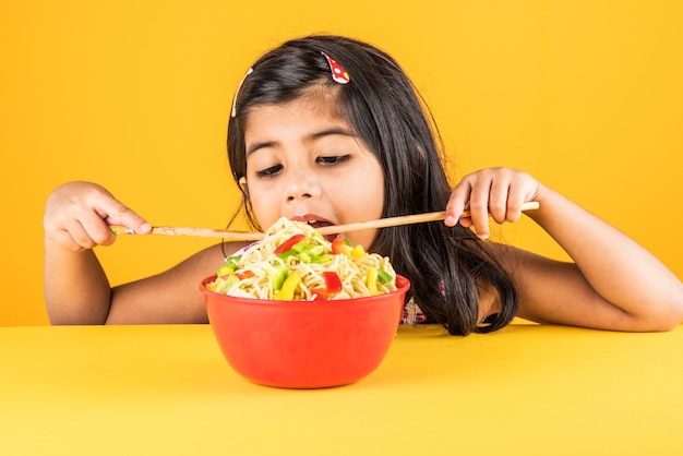 Fofinho menina indiana ou asiática comendo macarrão chinês saboroso com garfo ou pauzinhos, isolado sobre um fundo colorido