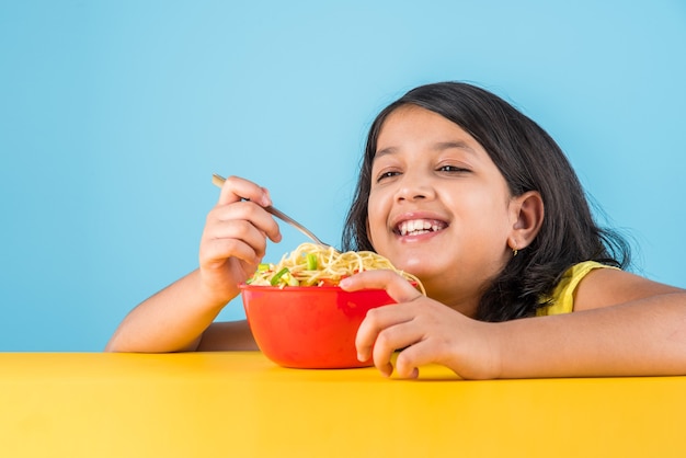 Fofinho menina indiana ou asiática comendo macarrão chinês saboroso com garfo ou pauzinhos, isolado sobre um fundo colorido