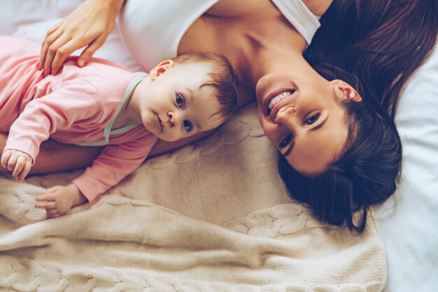 Fofinhos alegres. Vista superior de uma jovem bonita e alegre olhando para a câmera com um sorriso enquanto estava deitada na cama com sua filha