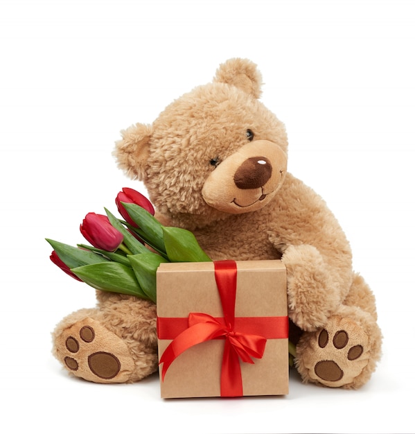 Fofo urso de pelúcia marrom tem na pata um buquê de tulipas vermelhas ao lado de um presente embrulhado
