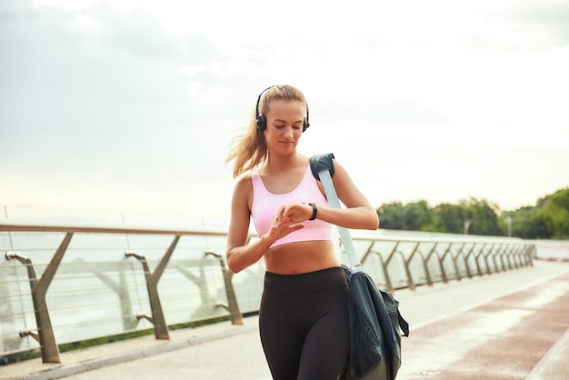 Foi ótimo treino, jovem mulher desportiva com fones de ouvido, carregando a bolsa e verificando o tempo