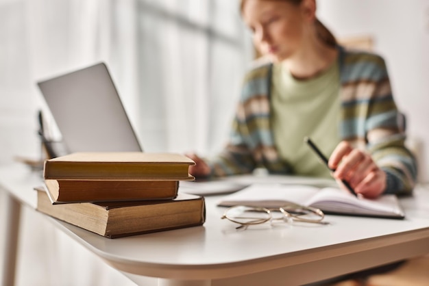 Fokus auf Bücher auf dem Schreibtisch Teenagerin hält Stift und schaut auf Laptop, während sie von zu Hause aus studiert