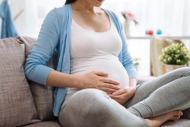 Fokus nah oben auf Bauch. junge elegante asiatische schwangere frau unerkannte hände, die kind im körper berühren, der auf couch sitzt, entspannen sich tagsüber in freizeitkleidung zu hause. chinesisches mädchen bereit, mutter eltern zu sein