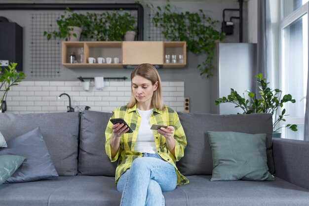 Fokussierte Frau beschließt, online einzukaufen. Ernsthafte Frau sitzt mit Telefon in der Hand und Bankkreditkarte auf dem Sofa