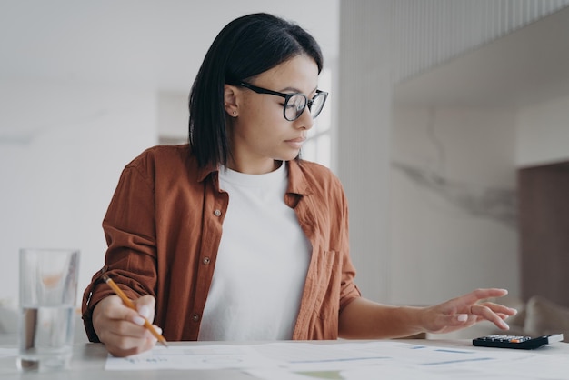 Fokussierte Frau mit Brille verwaltet die Ausgaben für die Budgetzählung auf dem Taschenrechner zu Hause Finanzmanagement