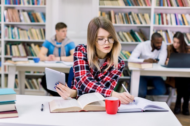 Fokussierte hübsche kaukasische Studentin, die im Coworking Space sitzt und mit Buch und Tablette studiert, Notizen macht und sich auf Test oder Prüfung in der Bibliothek vorbereitet