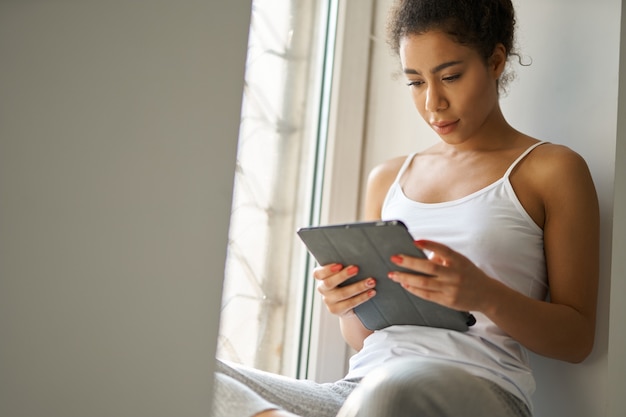 Fokussierte junge gemischtrassige Frau, die ein Buch mit Tablet-PC liest und sich beim Sitzen gemütlich fühlt