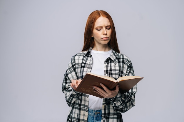 Fokussierte junge Studentin, die lässige Modekleidung trägt und Buch auf isoliertem grauem Hintergrund liest. Hübsches rothaariges Damenmodell, das emotional Gesichtsausdrücke im Studio zeigt, Kopierraum.