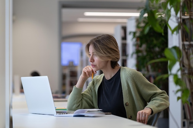 Fokussierte, nachdenkliche Hochschullehrerin, die sich auf einen Online-Vortrag auf einem Laptop in einer modernen Bibliothek vorbereitet