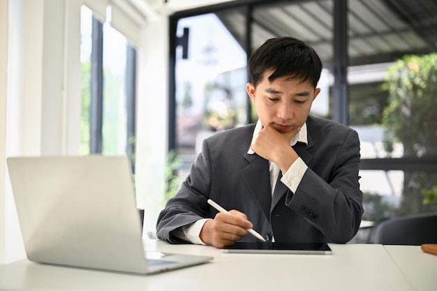 Fokussierter asiatischer Geschäftsmann mit seinem digitalen Tablet an seinem Schreibtisch im Büro