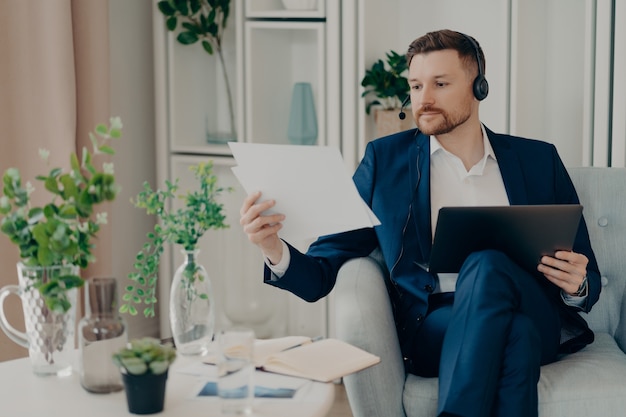 Fokussierter Geschäftsmann in Anzug und Headset, der im Sessel sitzt und den Laptop auf den Knien benutzt, Projektdokumente analysiert und über Geschäftsziele nachdenkt, während er zu Hause online spricht talking
