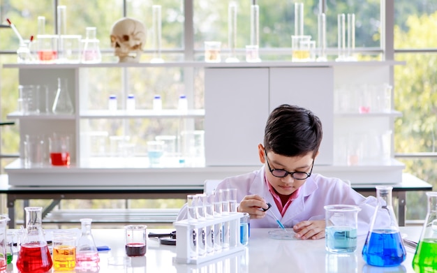 Fokussierter intelligenter asiatischer Schuljunge in Laborkittel und Brille, der Proben untersucht, während er Chemie in einem modernen Labor studiert.